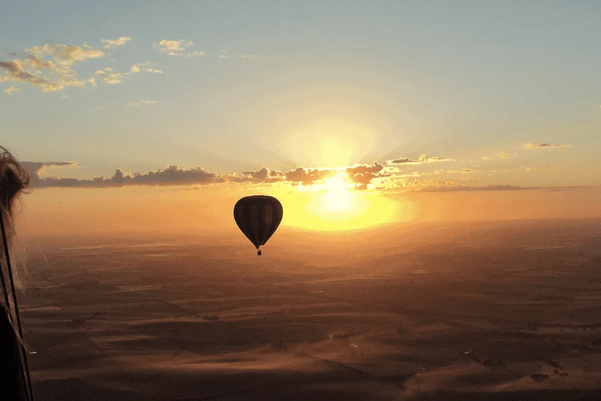 A silhouette of people in a hot air balloon, looking out at a sunrise and a hot air balloon in the distance