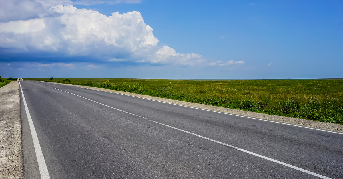 A long highway with no cars on the overgrown grass of the steppe under a blue cloudy sky